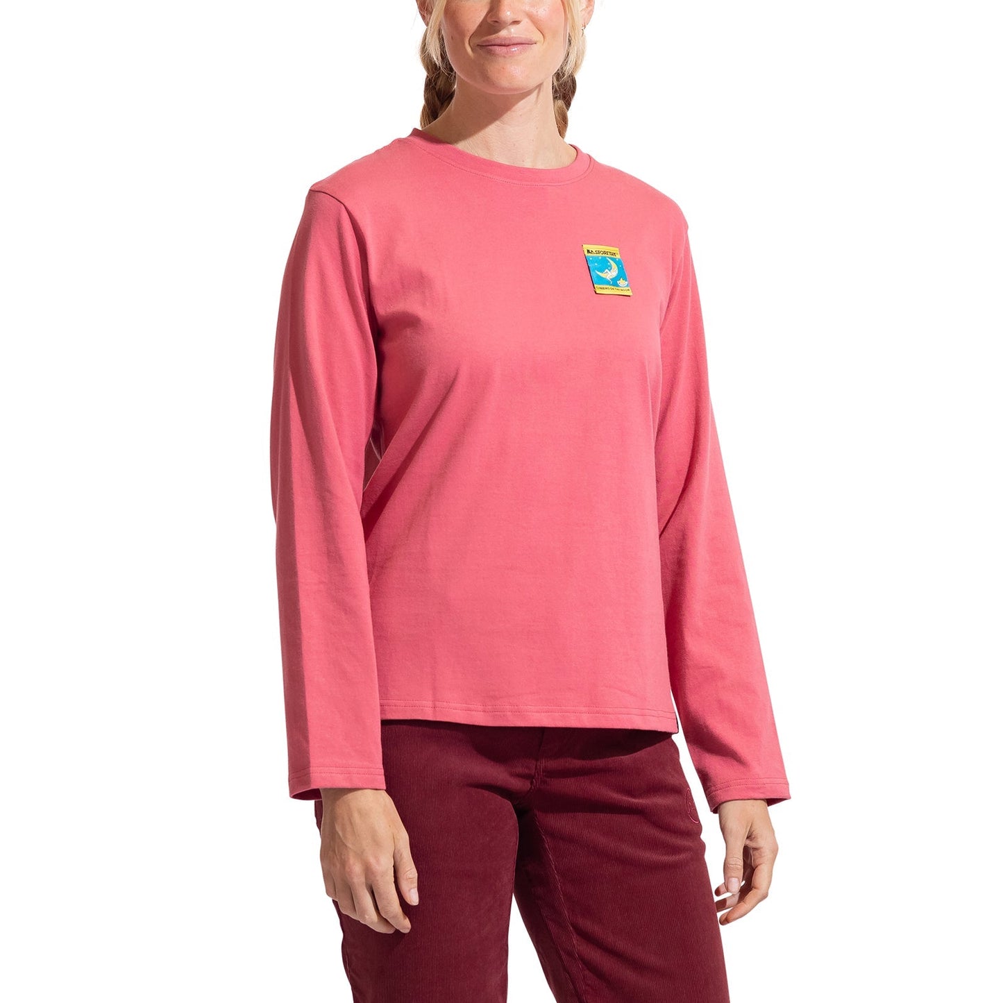 A person wearing the La Sportiva Moon Climb Long Sleeve Women's shirt in pink with a small colorful patch on the upper left chest and dark red pants stands against a white background.