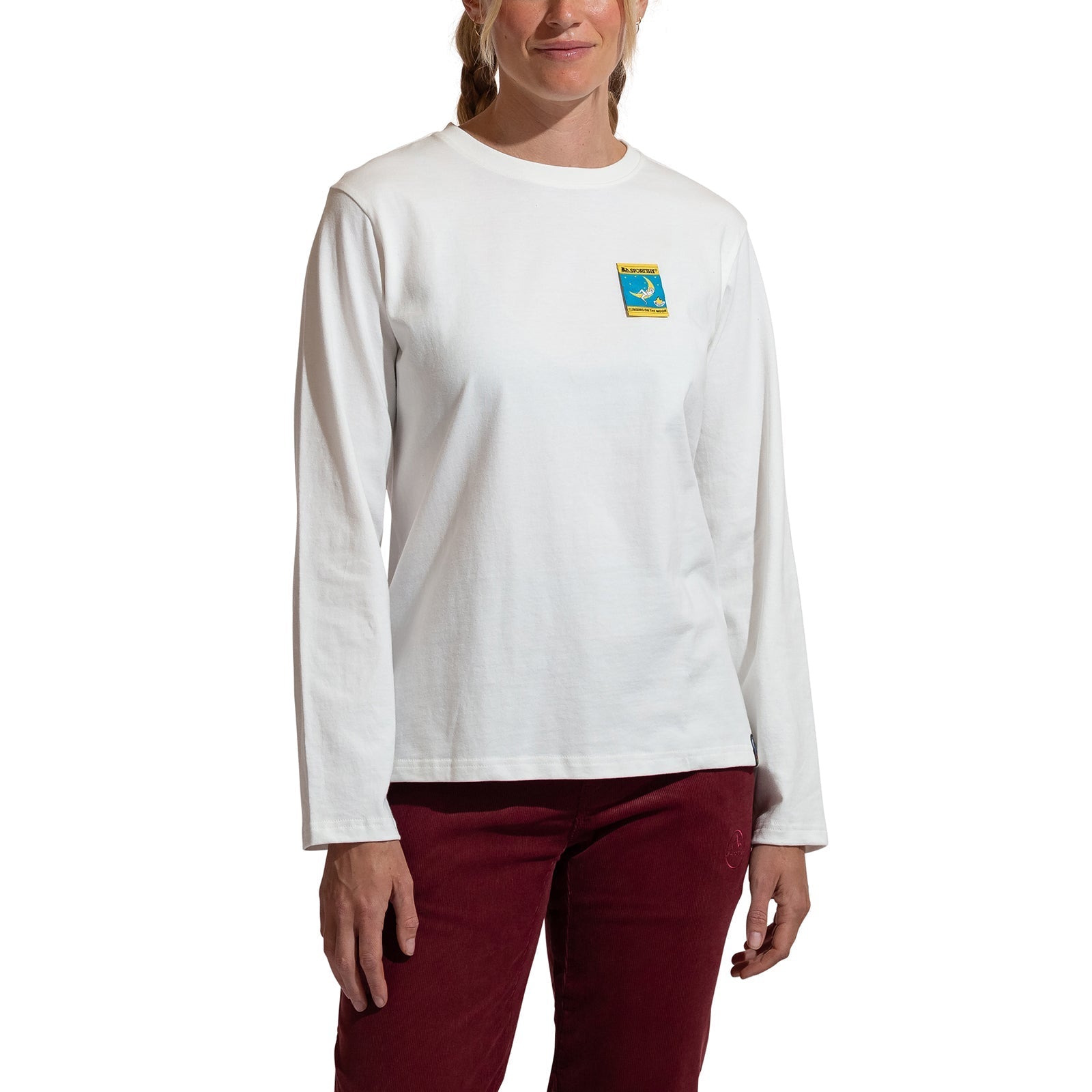 A woman models the La Sportiva Moon Climb Long Sleeve Women's shirt, featuring an organic cotton longsleeve design with a colorful chest patch, paired with maroon pants, against a plain white background.