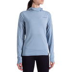 A woman wears the La Sportiva Cloud Shadow Sun Hoody—a lightweight, light blue UPF 50 long-sleeve with thumb holes—paired with black leggings, standing against a white background.
