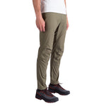 A man wears La Sportiva Roots Pants Men's in olive green, paired with a white tee and black hiking shoes with red laces. Only his lower body is shown against a plain white background in this minimalist design shot.