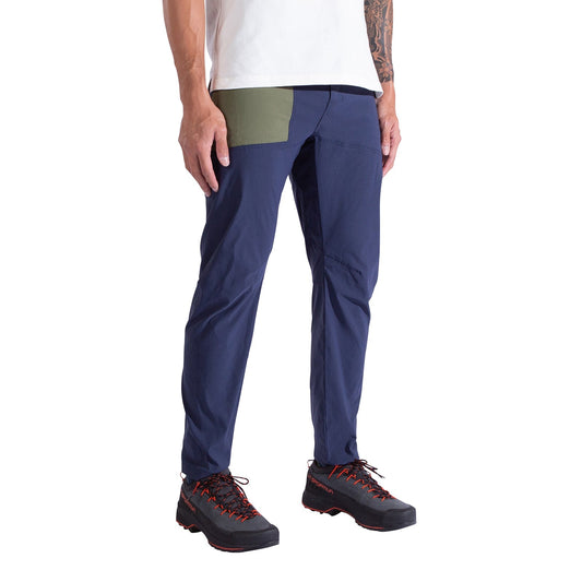 A person wearing La Sportiva Talus Pants Men's—eco-friendly blue climbing pants with a green thigh patch and articulated knees—as well as a white shirt and black hiking shoes with red laces. Only the lower half is visible.