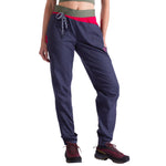 A woman wearing La Sportiva Bolt Pants in blue organic cotton with a green adjustable waistband, red accents, and drawstring, paired with black hiking shoes, stands against a white background.