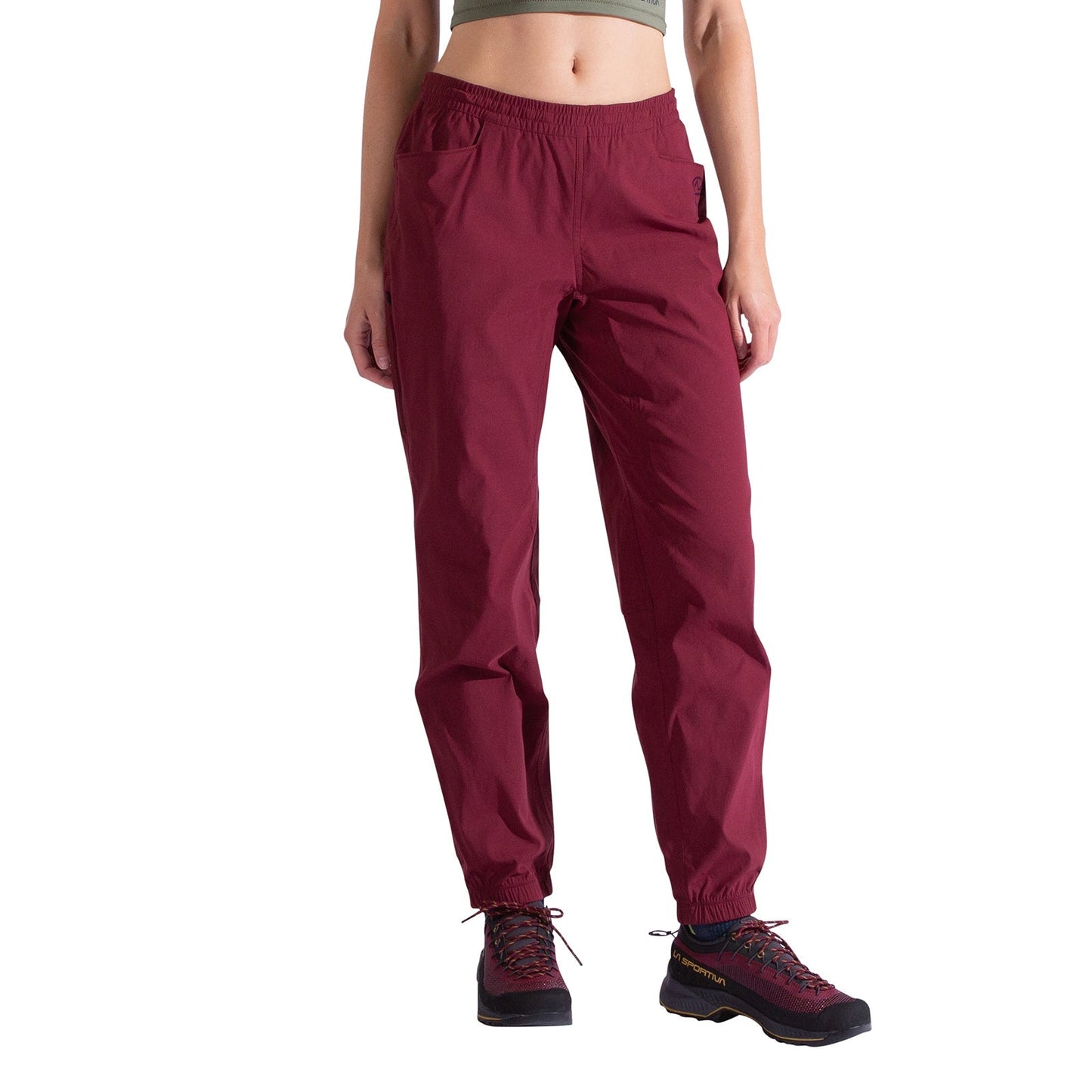 A person wears La Sportiva Gambit Pants Women's in burgundy, a green crop top, and black hiking shoes with red laces, standing against a white background with their head out of frame.