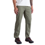 A person wearing La Sportiva Mainliner Pants Men's in olive green, a white t-shirt, and black hiking shoes with orange laces stands against a white background.