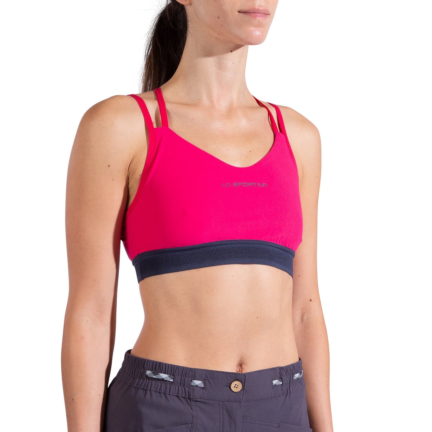 A woman models the La Sportiva Boulder Bra Women's in bright pink with double shoulder straps and dark gray high-waisted pants, standing against a white background.