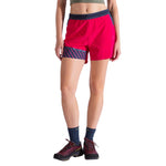 A person models La Sportiva Comp Shorts Women's—bright red with dark blue accents—alongside blue socks, hiking shoes, and a green crop top, standing against a white background. Their head is not shown in the image.