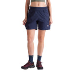 A person stands facing forward against a white background, wearing a green crop top, La Sportiva Roots Shorts Women's with durable pockets in navy blue, dark socks, and maroon hiking shoes.