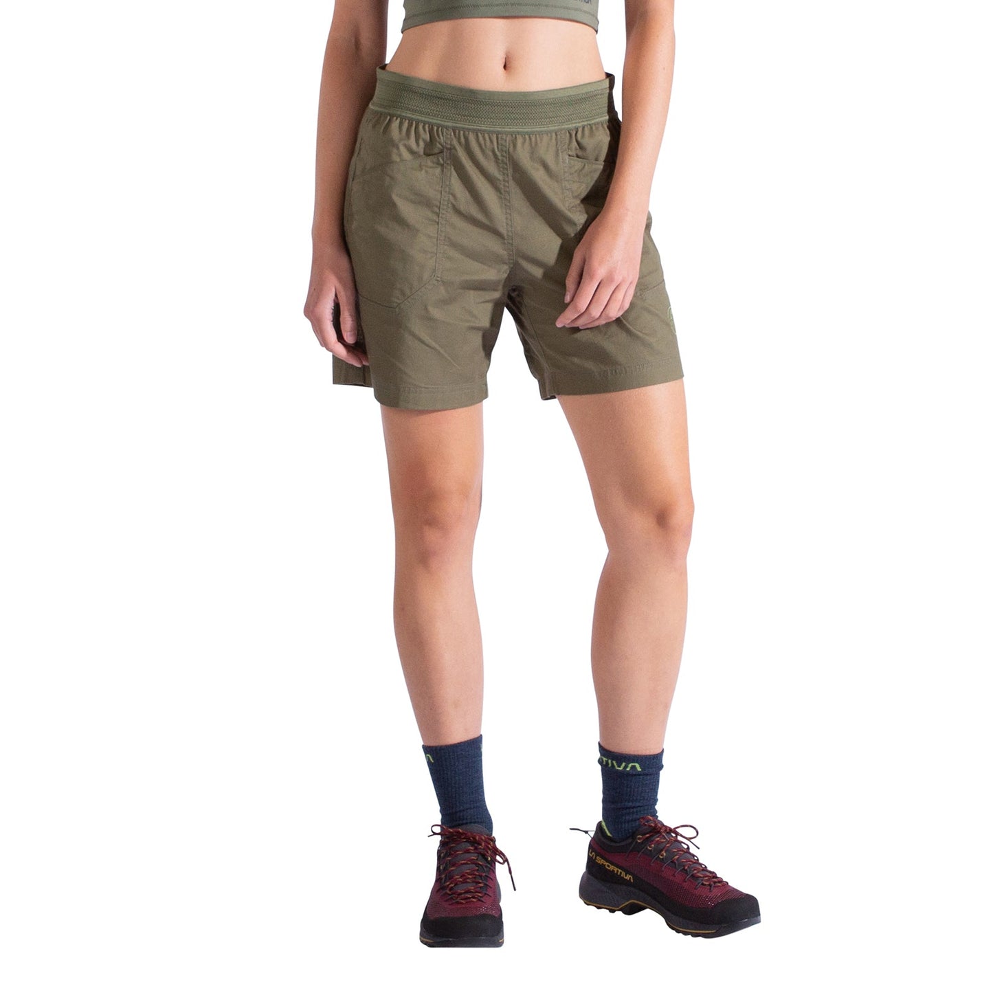 A woman wearing La Sportiva Roots Shorts in olive green with durable pockets, a matching crop top, navy socks, and maroon hiking shoes stands against a white background.