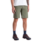 A person is shown from the waist down wearing La Sportiva Talus Shorts Men's in olive green, paired with a white T-shirt, navy socks, and black hiking shoes. The relaxed pose highlights the shorts' recycled fabric design.