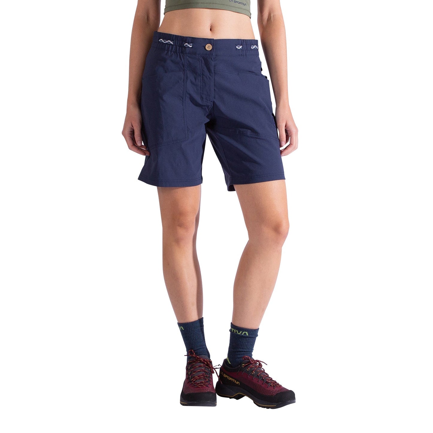 A person wears La Sportiva Mantra Shorts Women's in navy blue with a chalk bag loop, a cropped olive top, blue socks, and red-black hiking shoes. Their face is not visible; the background is white.