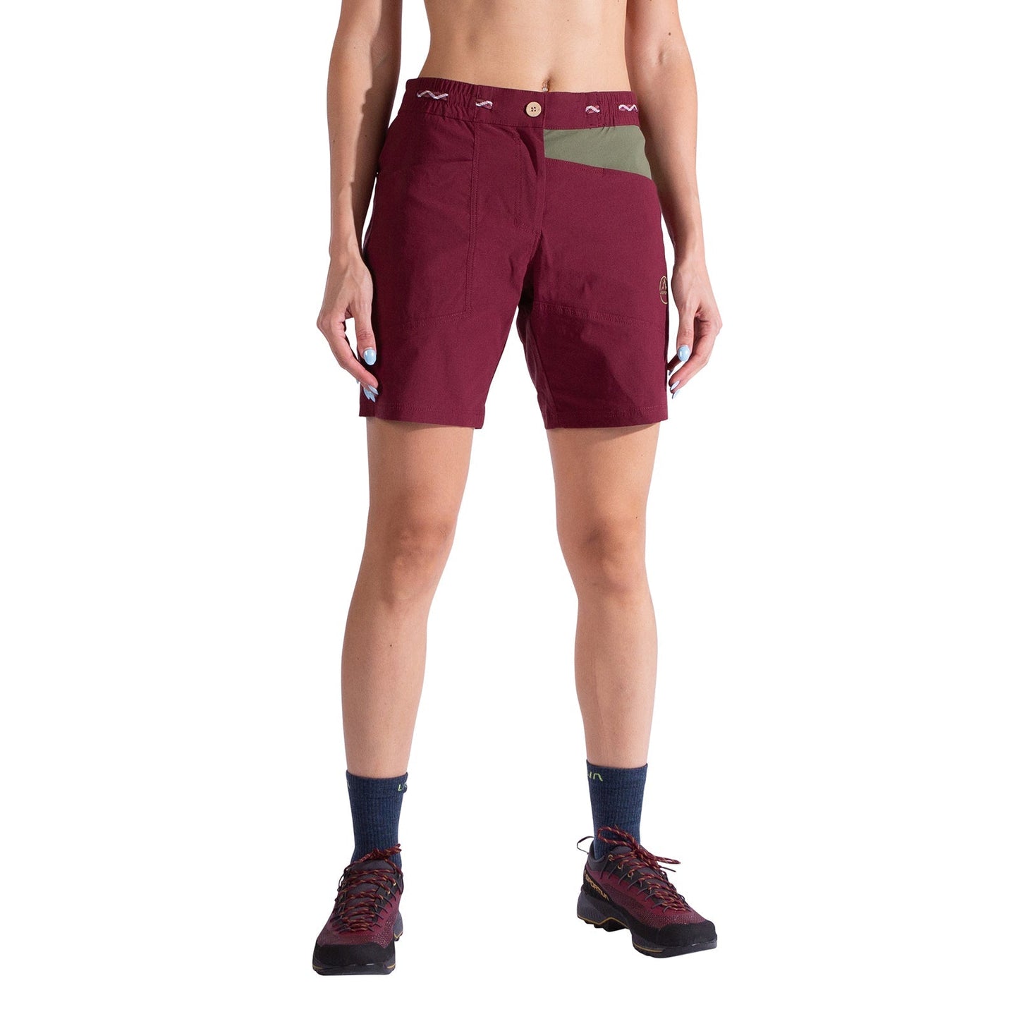 A woman stands against a white background wearing La Sportiva Mantra Shorts Women's in burgundy with an olive green panel, navy socks, and hiking shoes. The shorts have a button closure, visible waistband details, and a convenient chalk bag loop.