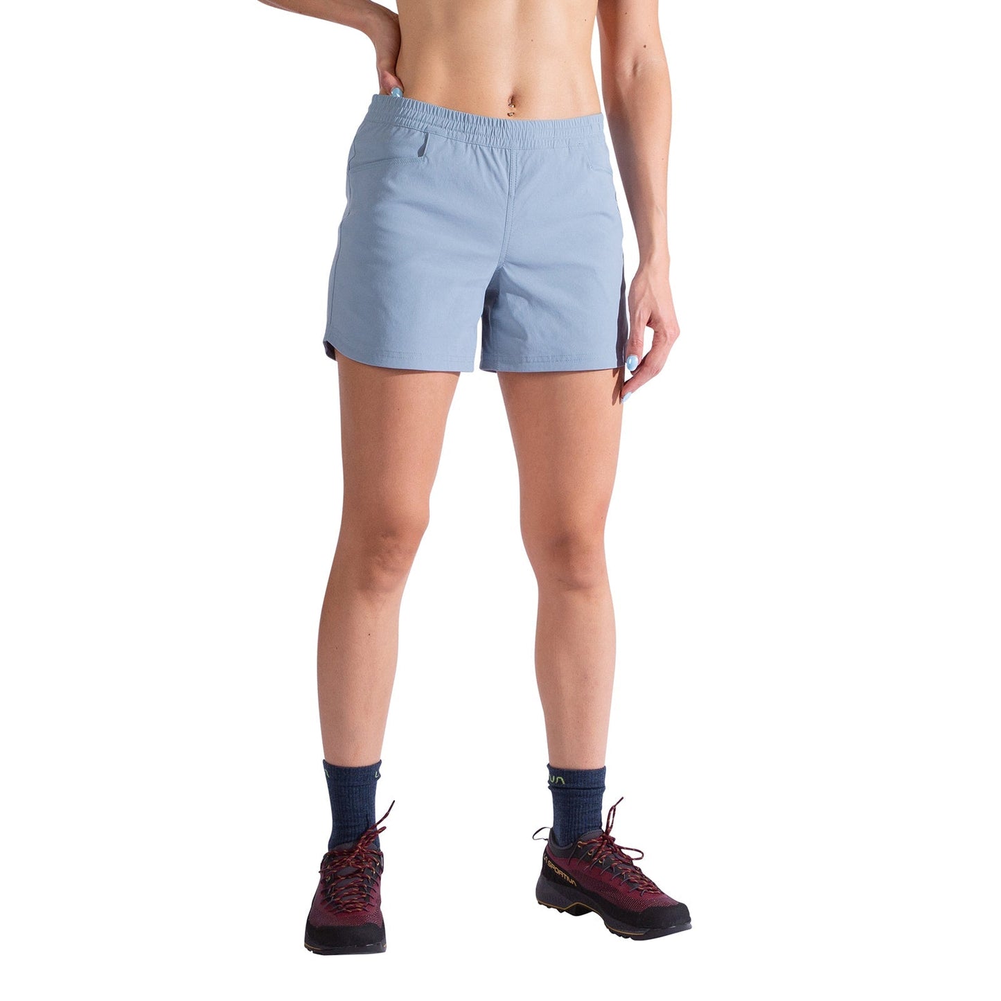 A person stands against a white background, showing only their torso and legs. They are wearing La Sportiva Gambit Shorts Women's in light blue, paired with a crop top, dark blue socks, and maroon hiking shoes.