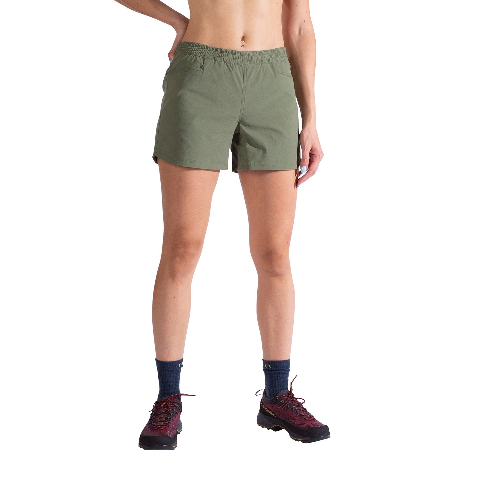 A person models La Sportiva's Gambit Shorts Women's in olive green, paired with a cropped top, navy blue socks, and dark hiking shoes. They stand with one hand on their hip against a white background; their head is not shown.