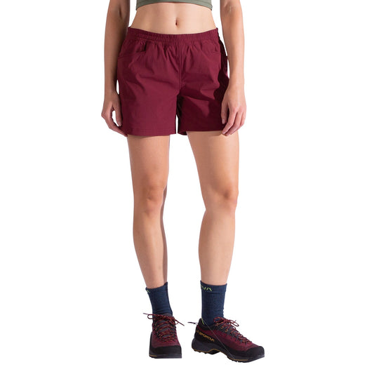 A woman wearing La Sportiva Gambit Shorts in burgundy, a green crop top, blue socks, and burgundy trail running shoes stands against a white background. The image shows her outfit from the neck down; her head is not visible.