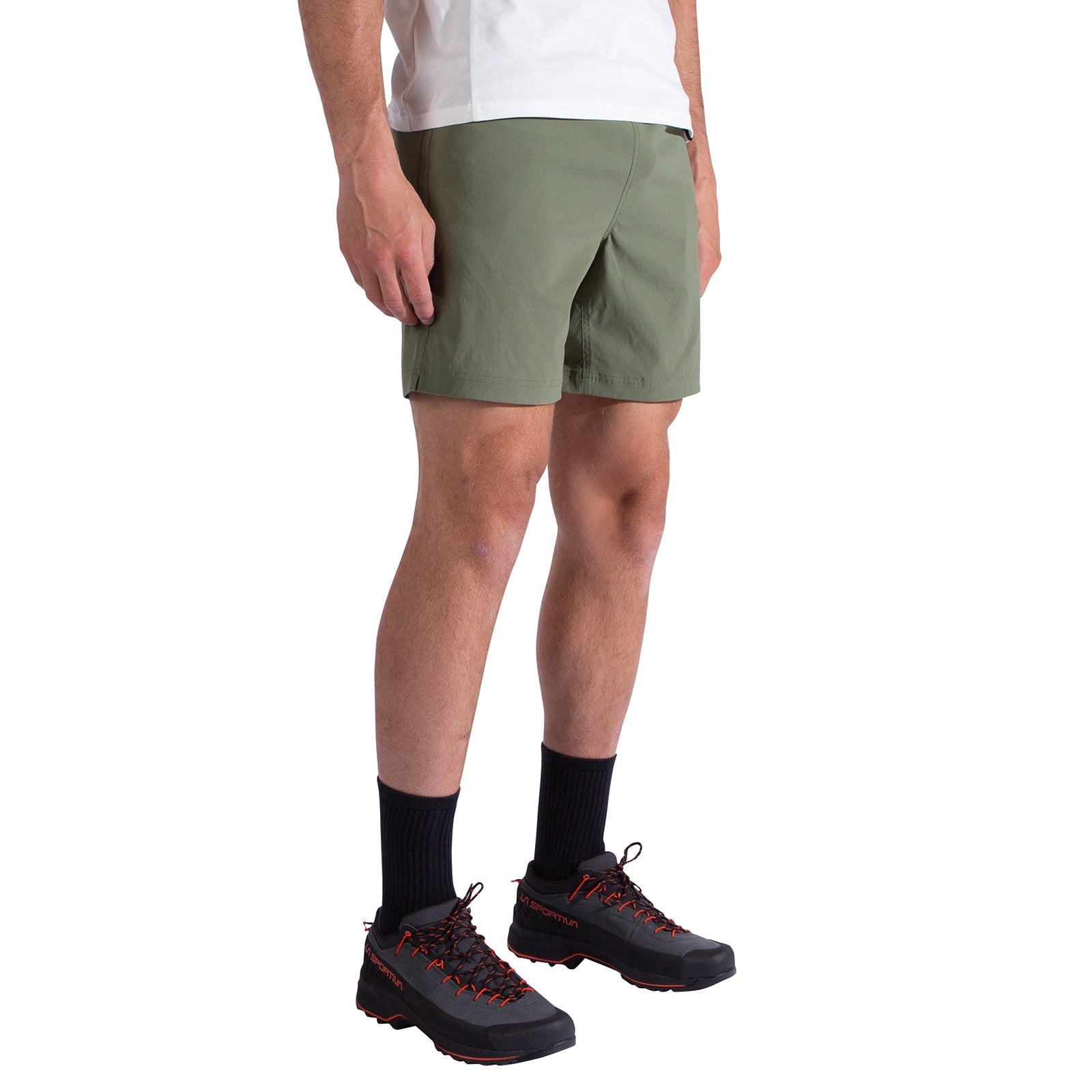 A person wearing olive green La Sportiva Gambit Shorts Men's with a white shirt, black socks, and black hiking shoes with red laces stands against a white background.