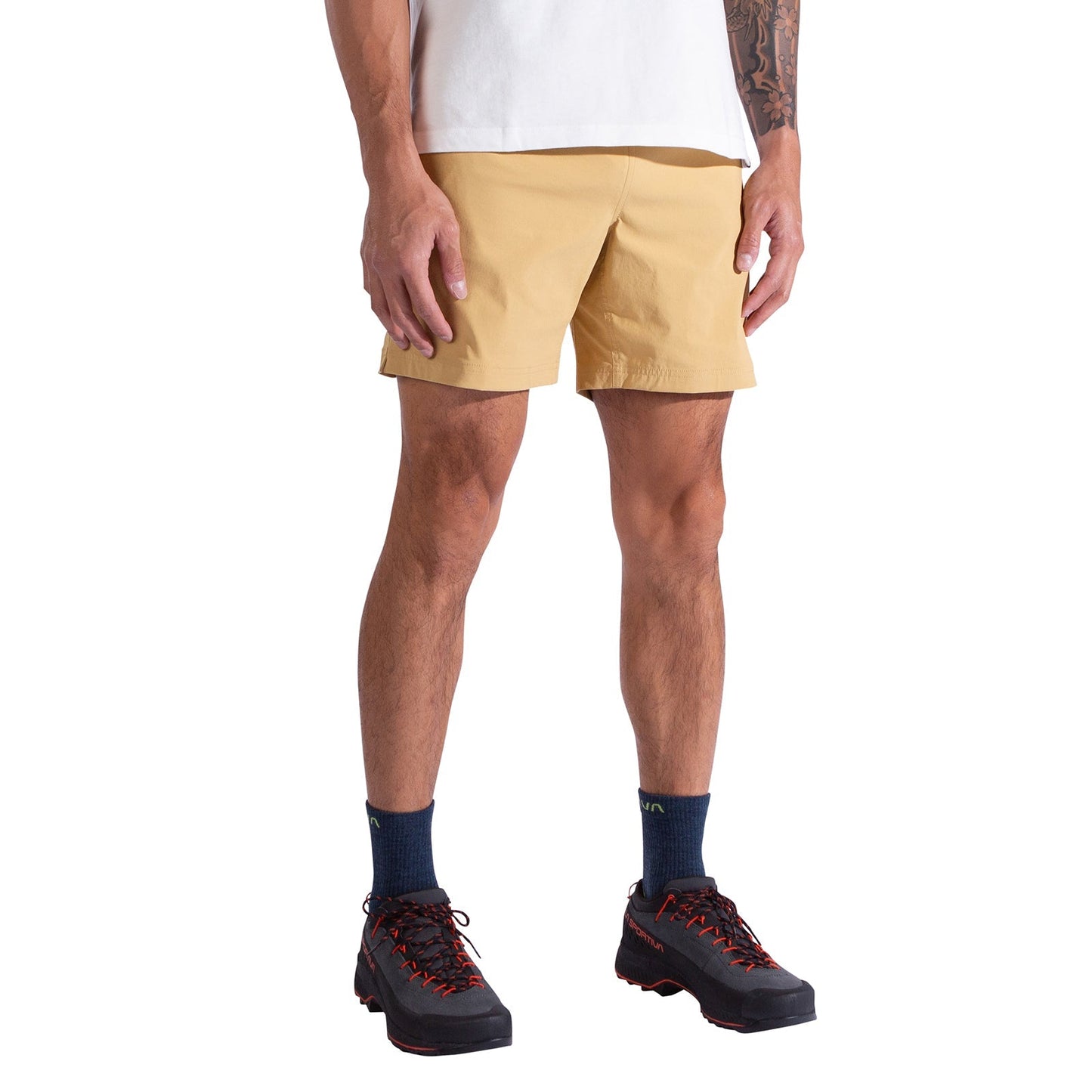 A person stands against a white background, arms relaxed at their sides, wearing a white shirt, La Sportiva Gambit Shorts Men's—lightweight, breathable hiking shorts—navy socks, and black trail shoes with orange laces.