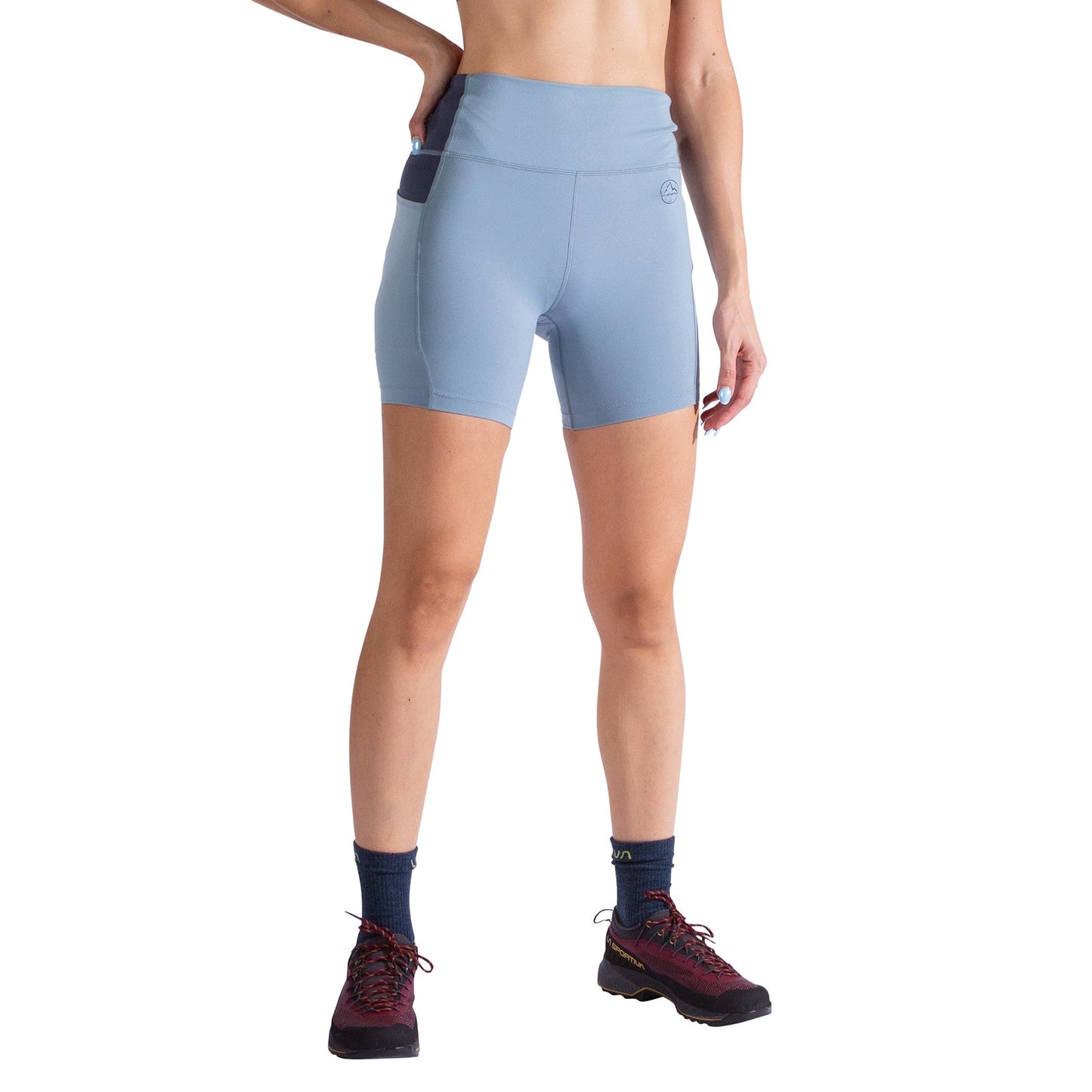 A person wears La Sportiva Cosmosis 6 Inch Shorts Women's in light blue with a side pocket, paired with dark socks and black athletic shoes. One hand is on their hip, and the image is cropped at the shoulders.