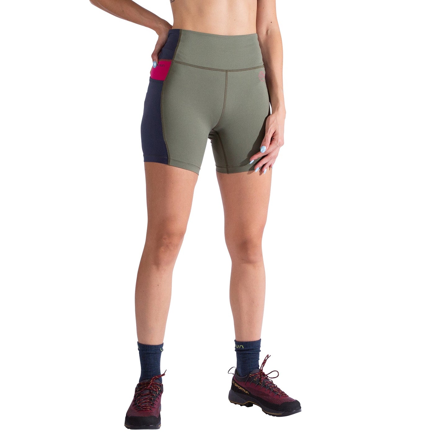 A person in La Sportiva Cosmosis 6 Inch Shorts Women's (green/navy with pink pocket), navy socks, and red trail shoes stands with one hand on their hip against a white background.