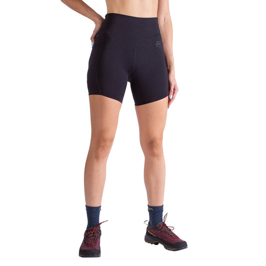 A woman wearing La Sportiva Cosmosis 6 Inch Shorts stands against a white background, blue socks and red-black trail shoes visible. Her upper body is partly out of frame.