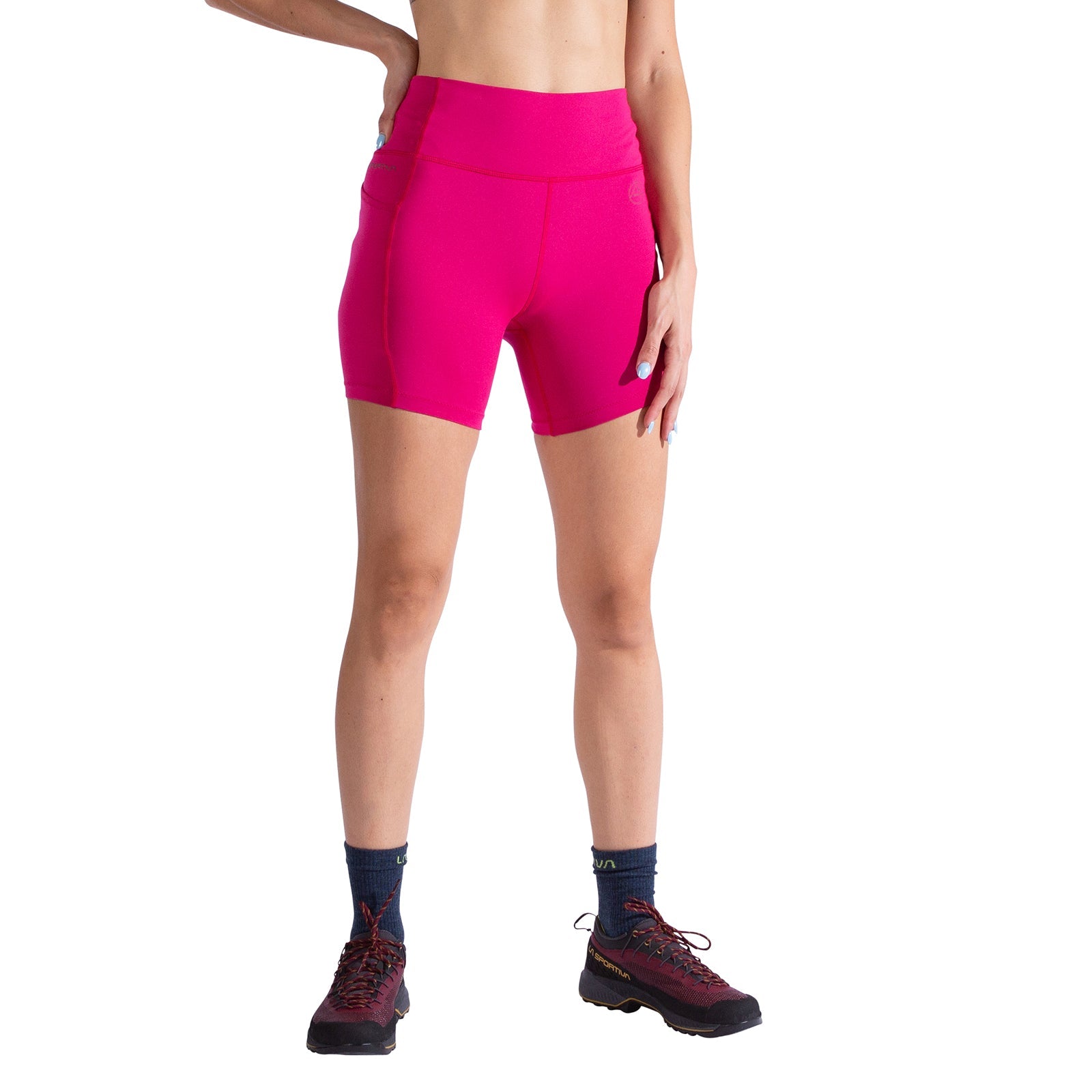 A person wearing La Sportiva Cosmosis 6 Inch Shorts Women's in bright pink, dark socks, and black trail running shoes stands with one hand on their hip against a white background. Their face is not shown.