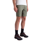 A person wears La Sportiva Mainliner Shorts Men's in olive green, paired with a white T-shirt, black socks, and black hiking shoes with red laces, standing against a plain white background.