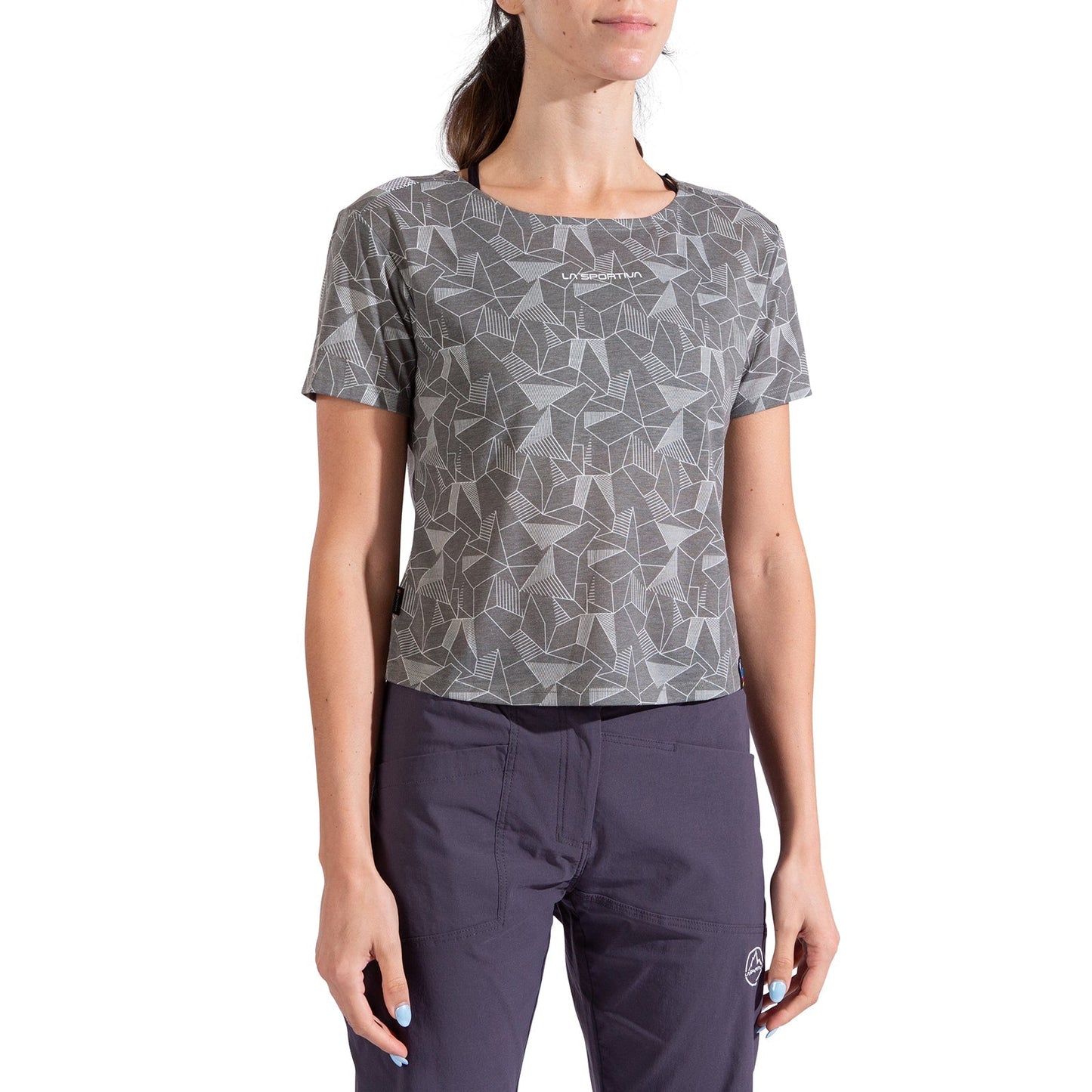A woman stands against a white background wearing the La Sportiva Dimension T-Shirt Women's, a gray geometric-patterned short-sleeve shirt made from recycled polyester with a subtle chest logo, paired with dark pants for an eco-friendly look.