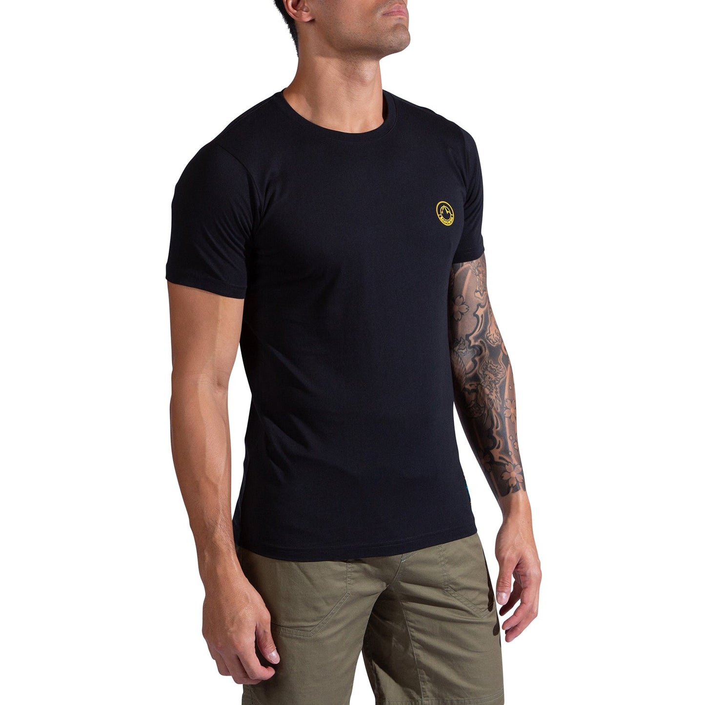 A man wearing the La Sportiva Climbing on the Moon T-Shirt in black, featuring a small yellow logo on the chest and paired with olive green pants. He stands against a white background, one tattooed arm visible, looking slightly upward.