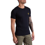 A man wearing the La Sportiva Climbing on the Moon T-Shirt in black, featuring a small yellow logo on the chest and paired with olive green pants. He stands against a white background, one tattooed arm visible, looking slightly upward.
