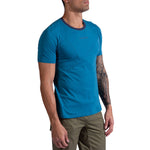 A man wearing the La Sportiva Dude T-Shirt Men's—a fitted blue tee with dark neck trim—and olive green pants stands sideways, showcasing a tattoo sleeve against a white background.