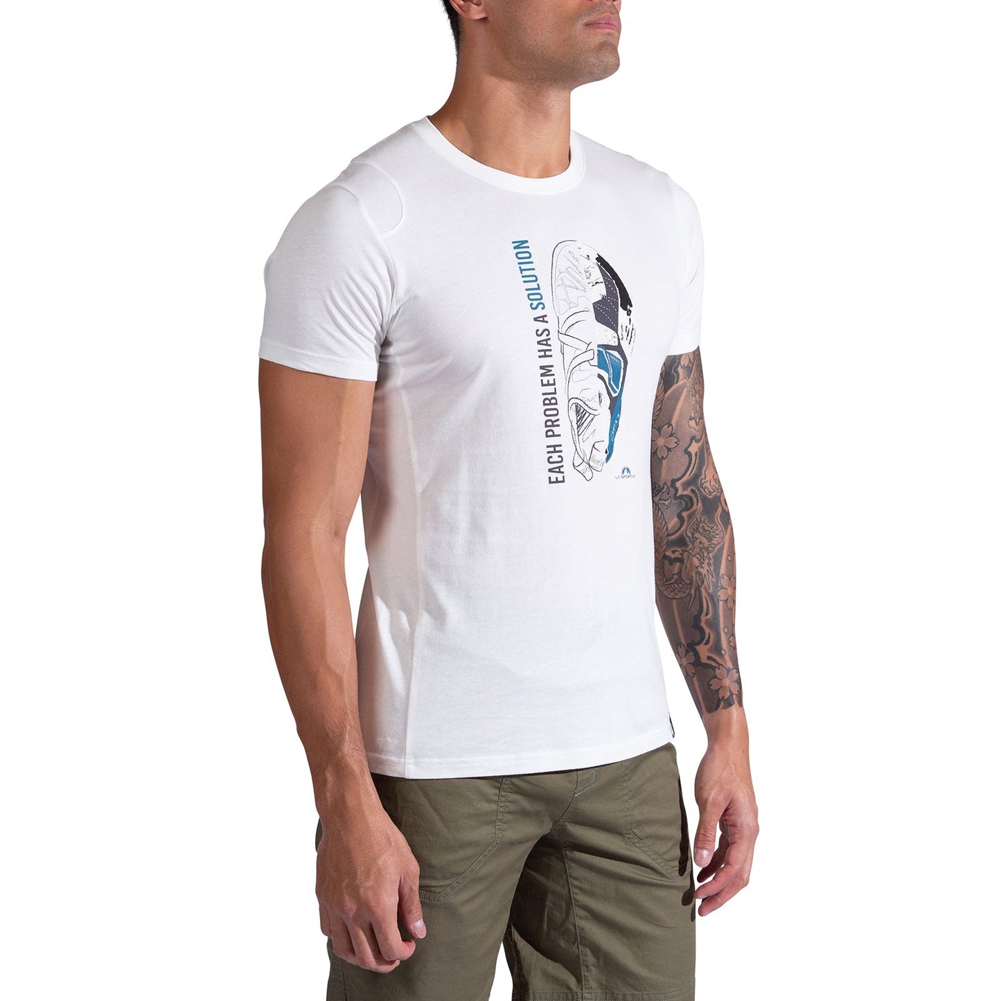 A man wears the La Sportiva Solution T-Shirt Men's, made from organic cotton with a climbing shoe graphic and "Each problem has a solution" text, paired with olive green pants. Ideal for climbers, his left arm displays a tattoo sleeve.