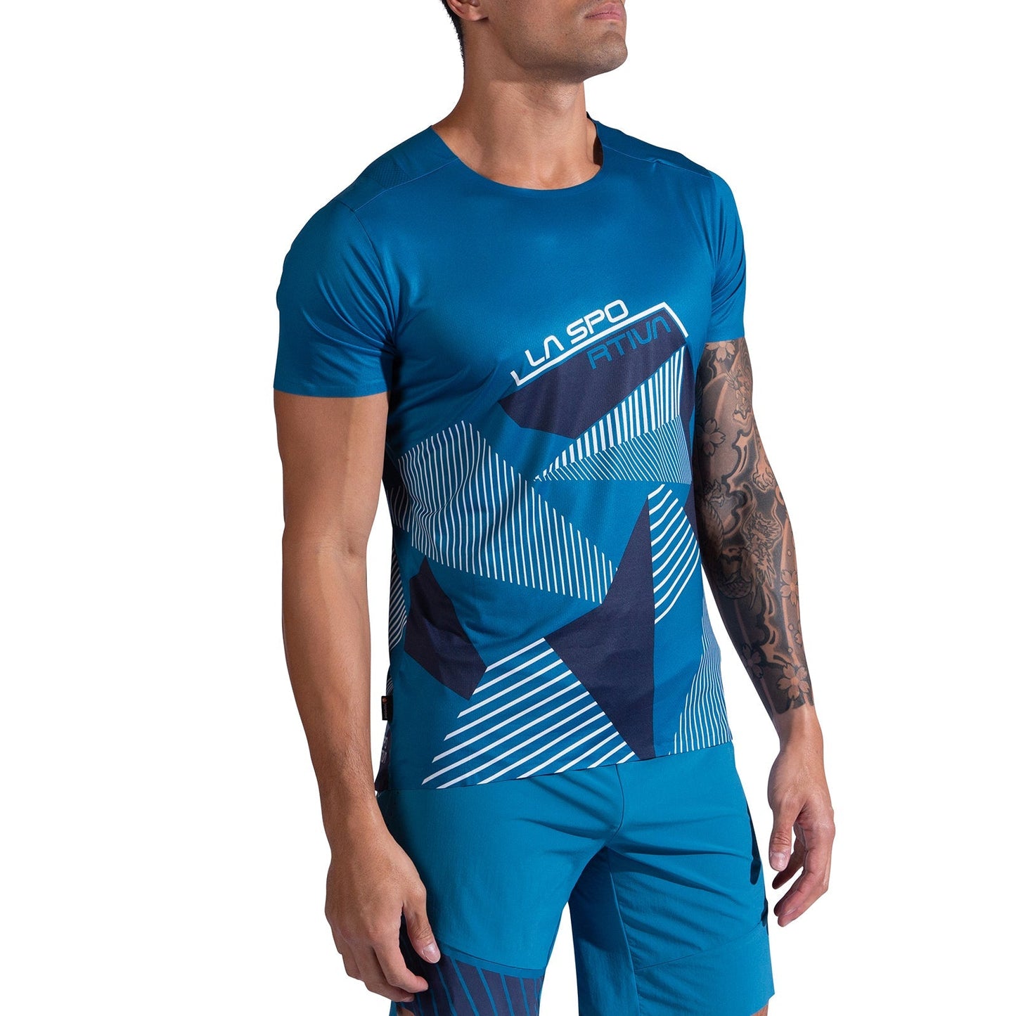 A man models the La Sportiva Comp T-Shirt Men's in blue with a geometric pattern, paired with matching shorts, standing with his right arm by his side and his tattooed left arm slightly bent.