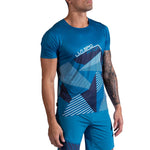 A man models the La Sportiva Comp T-Shirt Men's in blue with a geometric pattern, paired with matching shorts, standing with his right arm by his side and his tattooed left arm slightly bent.