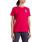 A person wearing the La Sportiva Moon Climb T-Shirt Women's, a bright pink organic cotton tee with a colorful left chest patch, and dark pants stands against a white background.