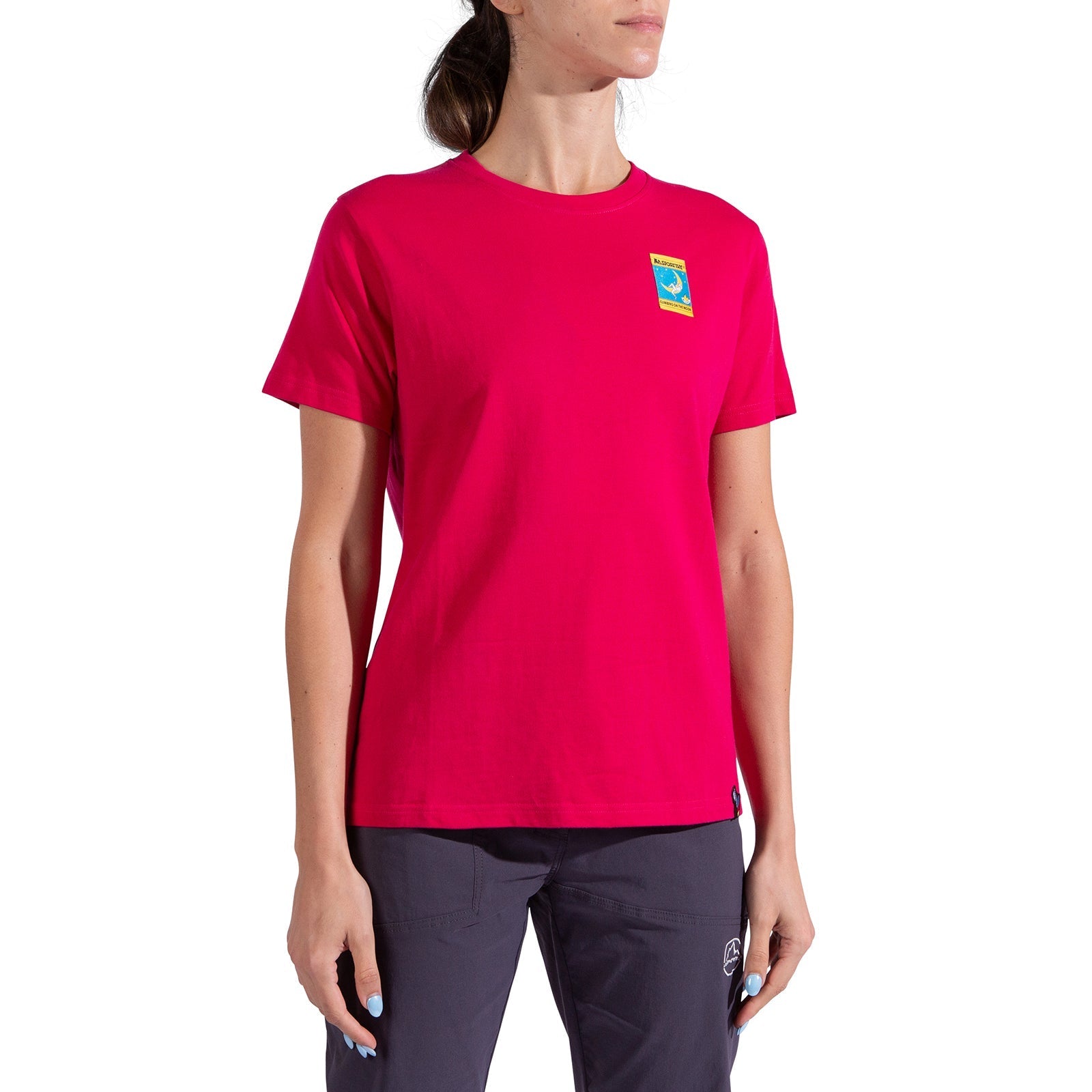 A person wearing the La Sportiva Moon Climb T-Shirt Women's, a bright pink organic cotton tee with a colorful left chest patch, and dark pants stands against a white background.