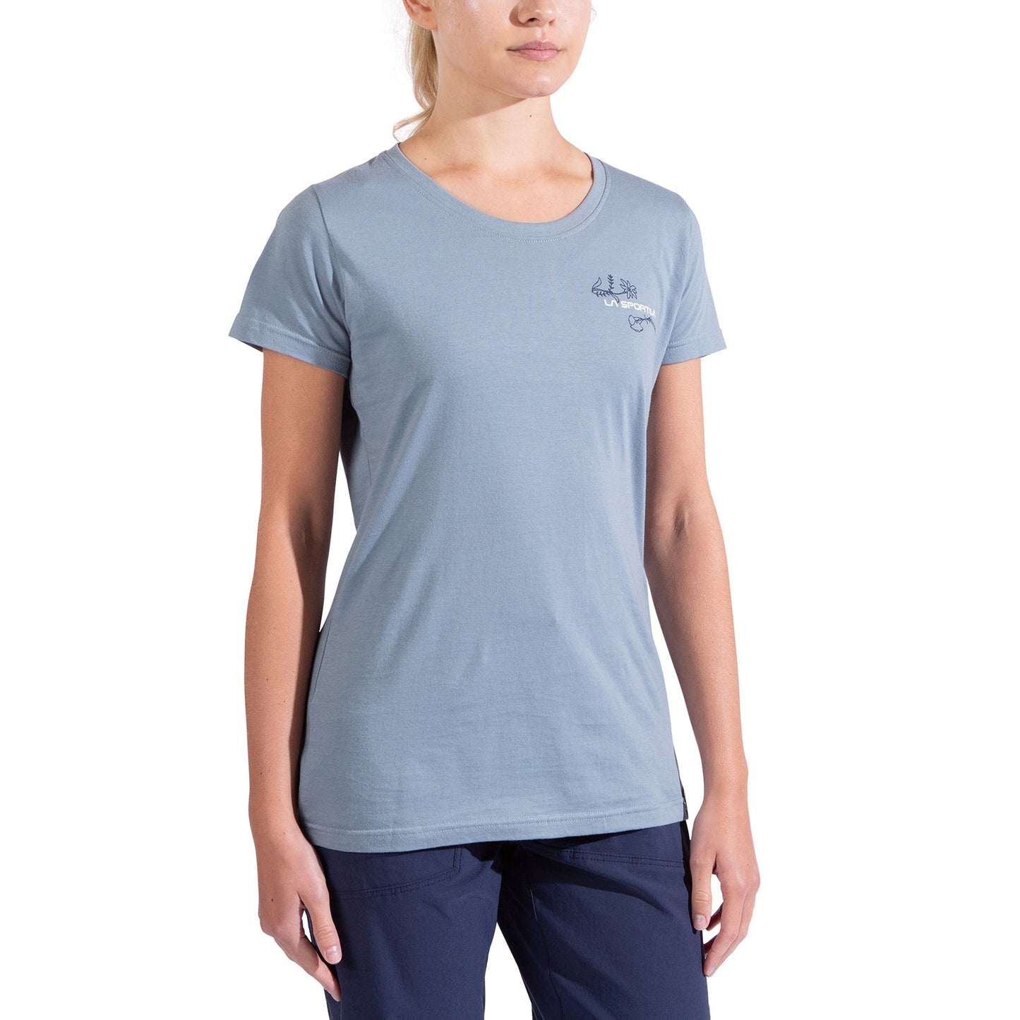 A woman in a light blue La Sportiva Climb Flower T-Shirt Women's, made from organic cotton with a small left-chest logo, and dark blue pants stands against a white background. Her face is partially out of frame.