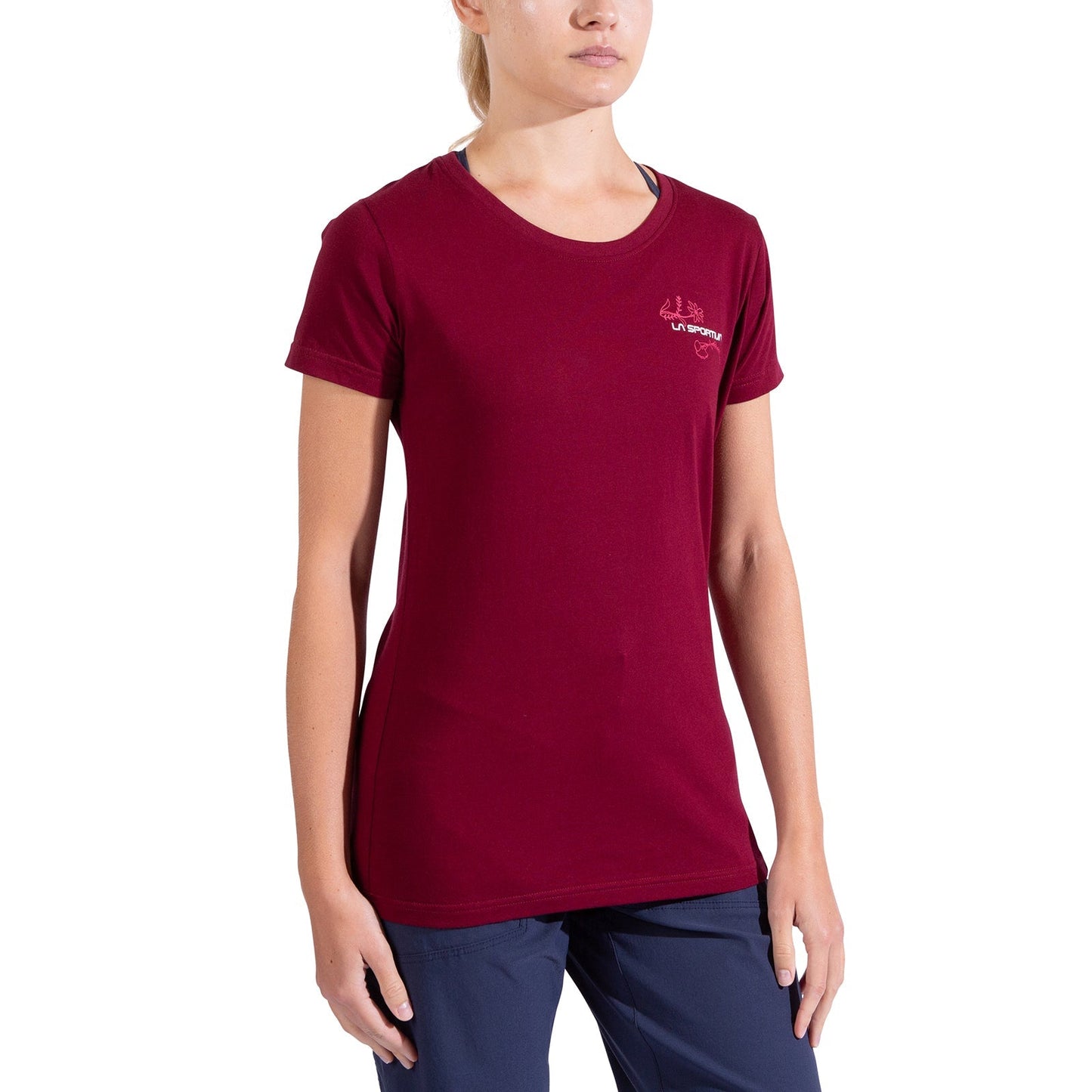 A woman wears a maroon La Sportiva Climb Flower T-Shirt Women's with a small white chest logo and navy pants, standing against a plain white background. Her head is partially out of frame.