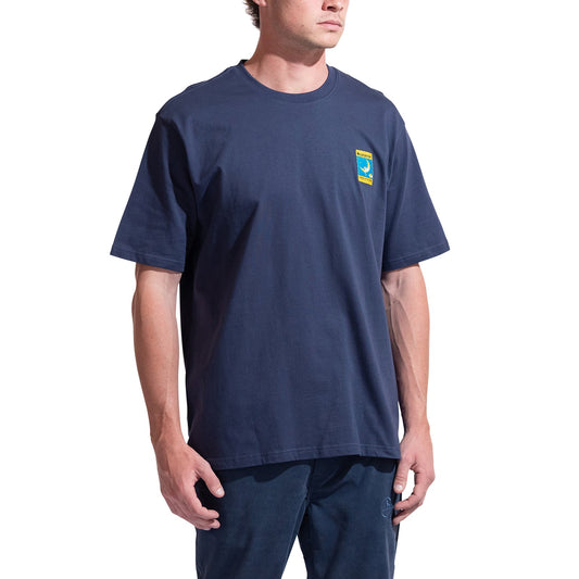 A person wears the La Sportiva Moon Patch T-Shirt Men's—a loose-fitting, organic cotton navy tee with a small, colorful patch on the left chest—paired with dark blue climber-friendly pants, standing against a white background.