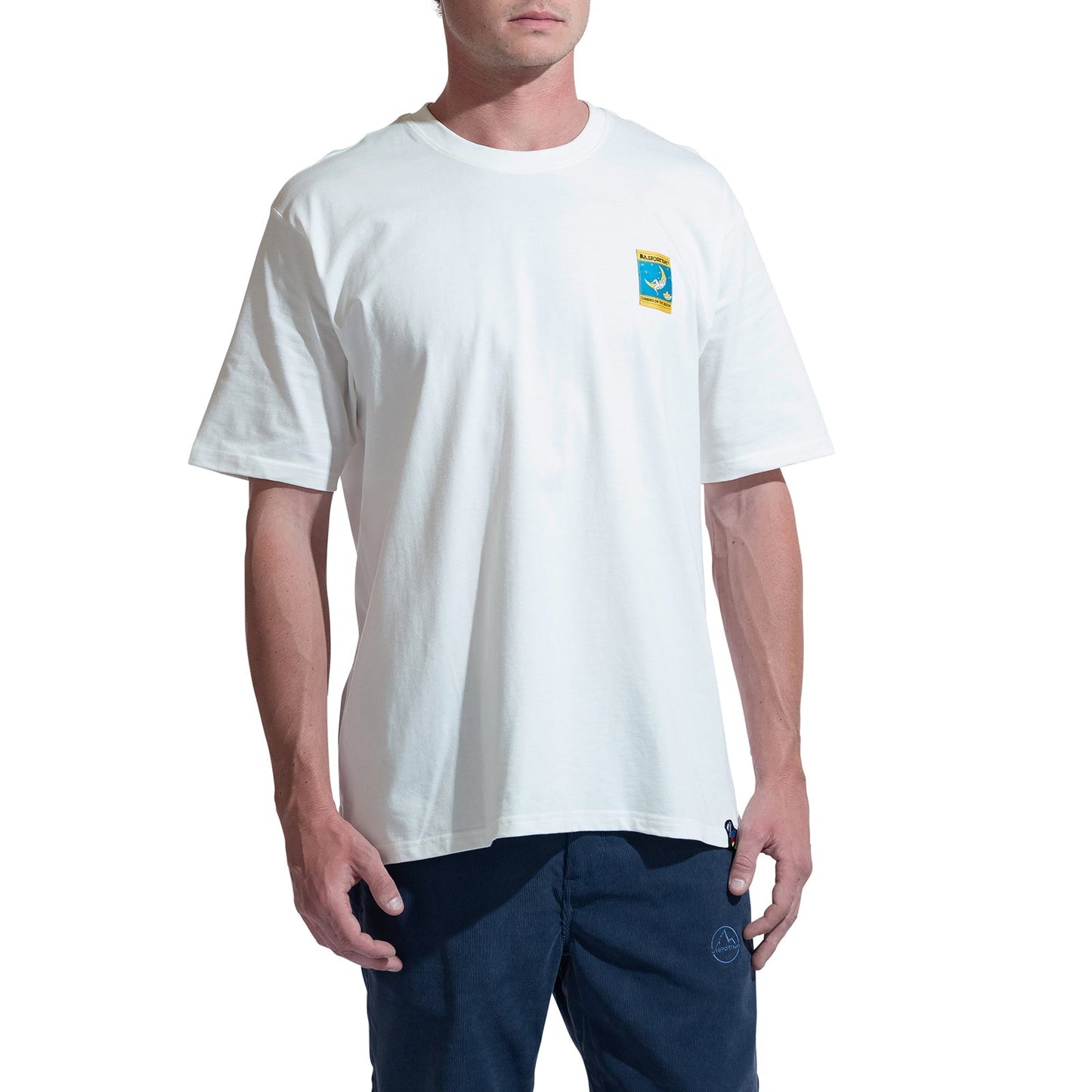 A person in the La Sportiva Moon Patch T-Shirt Men's—an oversized, organic cotton white tee with a colorful left-chest patch—and dark blue pants stands against a white background. Ideal for climbers who value comfort and style.