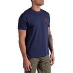 A man with a tattoo sleeve stands against a white background wearing La Sportiva's Circle Logo T-Shirt for men in navy blue, featuring a small red logo on the chest, paired with olive green shorts.