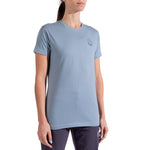 A woman with dark hair pulled back wears the La Sportiva Circle Logo T-Shirt Women's—a light blue organic cotton tee with a small circular logo on the left chest—styled with dark pants against a white background. Perfect for minimalist climbers.