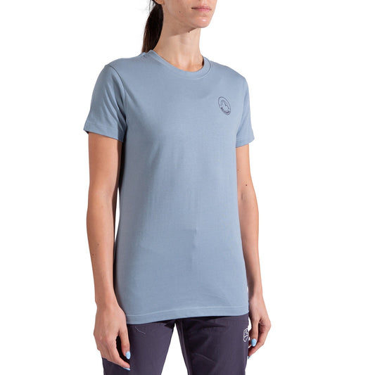 A woman with dark hair pulled back wears the La Sportiva Circle Logo T-Shirt Women's—a light blue organic cotton tee with a small circular logo on the left chest—styled with dark pants against a white background. Perfect for minimalist climbers.