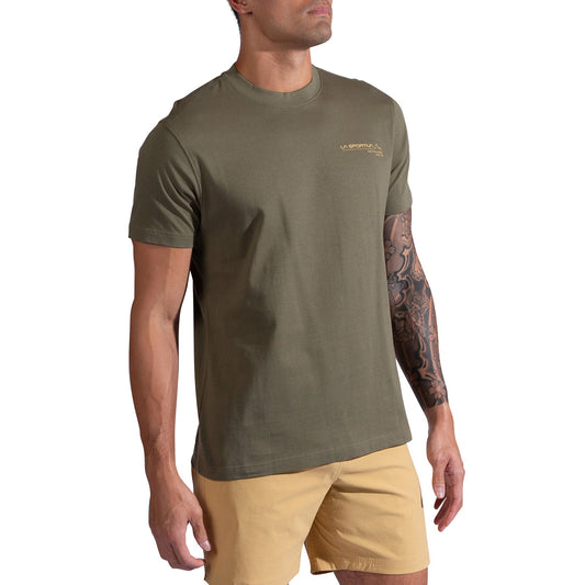 A man stands against a white background in a La Sportiva Since 1928 T-Shirt Men's in olive green, featuring a small chest logo, paired with tan shorts. He has a tattoo sleeve on his left arm.
