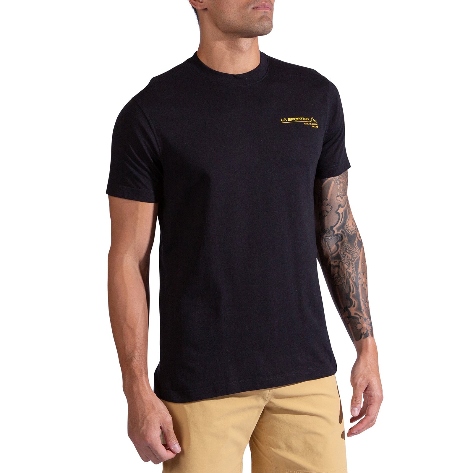A man with tattoos on his right forearm stands against a white background wearing the La Sportiva Since 1928 T-Shirt Men's in black, which features a small yellow logo on the chest, paired with tan shorts.
