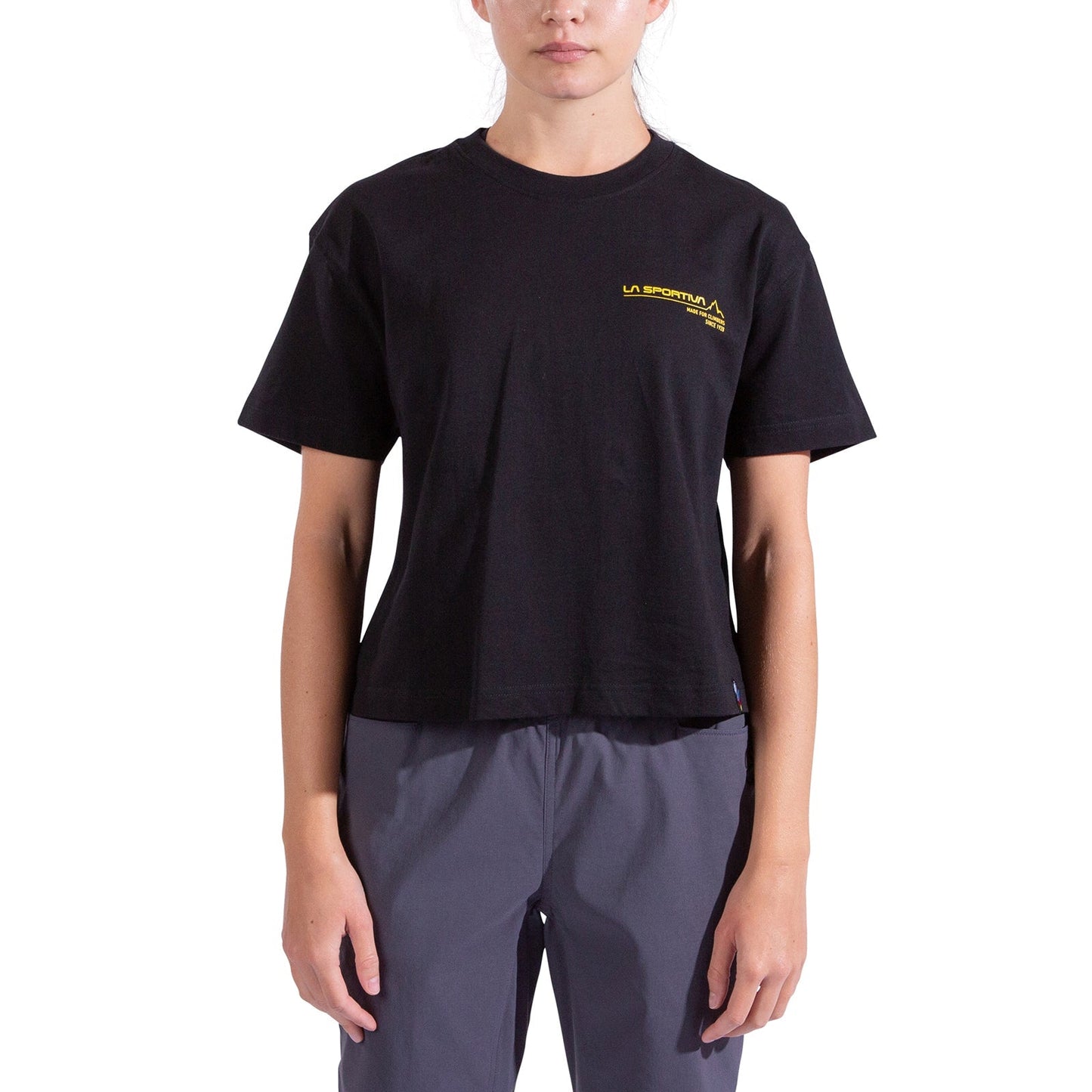 A woman wears the La Sportiva Since 1928 Crop T-Shirt in black, featuring a yellow logo on the left chest, paired with gray pants against a white background—ideal for climbing or everyday style.