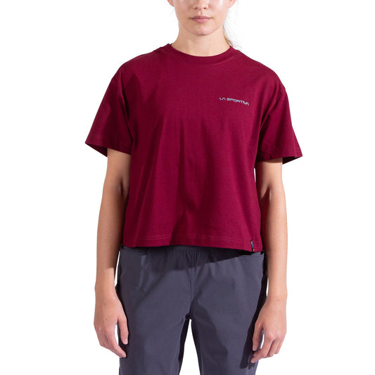 A woman wears the La Sportiva Trilogy Crop T-Shirt, a loose-fitting, heavyweight maroon organic cotton tee with branding on the left chest, paired with dark gray pants, standing against a white background.