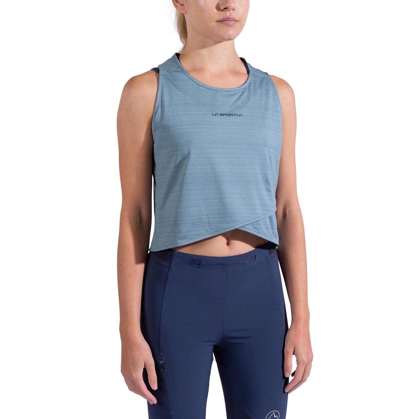 A woman wearing the La Sportiva Sunfire Tank Women's, a sleeveless, light blue cropped top, and dark blue high-waisted athletic pants stands against a white background.