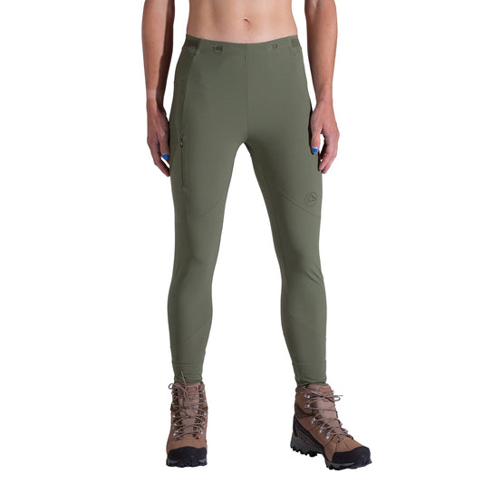 A woman faces forward against a white background, wearing La Sportiva Camino Tight Pants Women's in olive green with a zippered right thigh pocket and durable fabric, paired with brown hiking boots.
