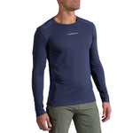 A man wears the La Sportiva Beyond Long Sleeve Men's top in dark blue, featuring moisture-wicking Polartec Delta Cooling Fabric, paired with olive green pants, standing against a plain white background.