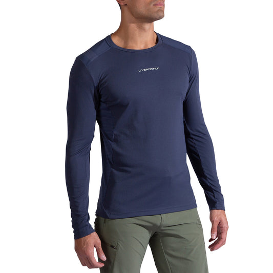 A man wears the La Sportiva Beyond Long Sleeve Men's top in dark blue, featuring moisture-wicking Polartec Delta Cooling Fabric, paired with olive green pants, standing against a plain white background.