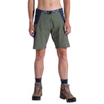 A woman wearing La Sportiva West Crest Shorts in olive green and navy, navy socks, and brown hiking boots stands facing forward against a white background. Only her body from the waist to feet is visible.